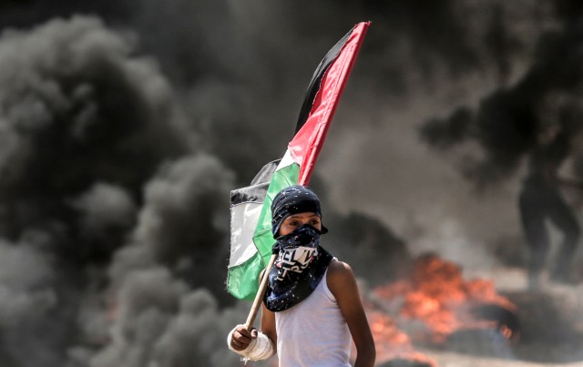 Un niño palestino sosteniendo su bandera nacional observa los enfrentamientos con las fuerzas de seguridad israelíes cerca de la frontera entre Gaza e Israel al este de la ciudad de Gaza el 14 de mayo de 2018, mientras los palestinos protestan por la inauguración de la embajada de Estados Unidos tras su controvertido traslado a Jerusalén. Docenas de palestinos fueron asesinados por disparos israelíes el 14 de mayo cuando decenas de miles protestaron y estallaron enfrentamientos a lo largo de la frontera de Gaza contra la transferencia de su embajada a Jerusalén, luego de meses de protestas globales, ira palestina y elogios exuberantes de los israelíes por el presidente Donald Trump decisión dejando de lado décadas de precedentes. / AFP PHOTO / MAHMUD HAMS